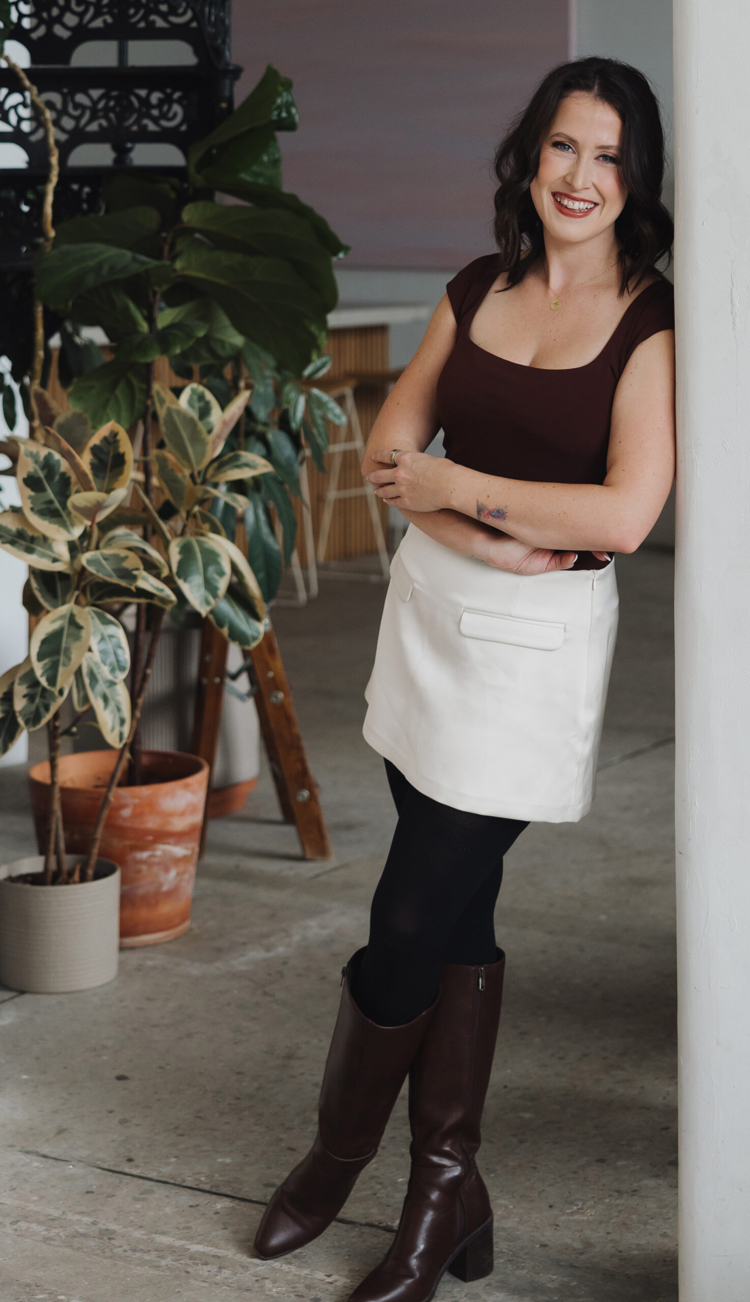 Woman with shoulder-length dark hair, wearing a brown top, white skirt, black tights, and brown boots, leans against a white column in a modern indoor space with plants and contemporary decor—an elegant setting ideal for a photo shoot or professional headshots.