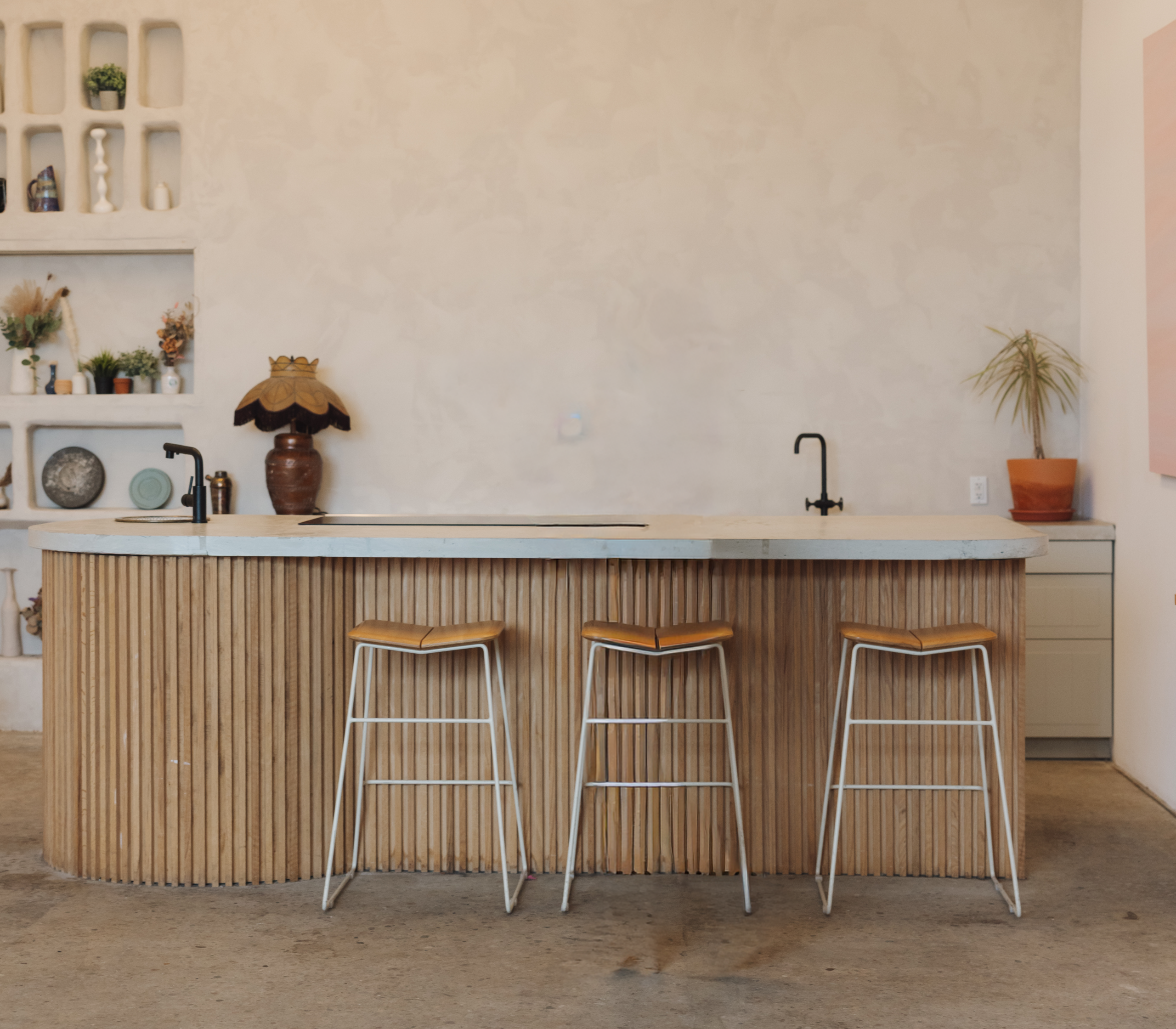 Modern kitchen with a curved wooden island, three bar stools, two black faucets, open shelves displaying decor, and neutral tones throughout. Plants and pottery add a touch of style to the minimalist space—perfectly highlighted through real estate photography or enhanced by virtual staging for an elevated presentation.