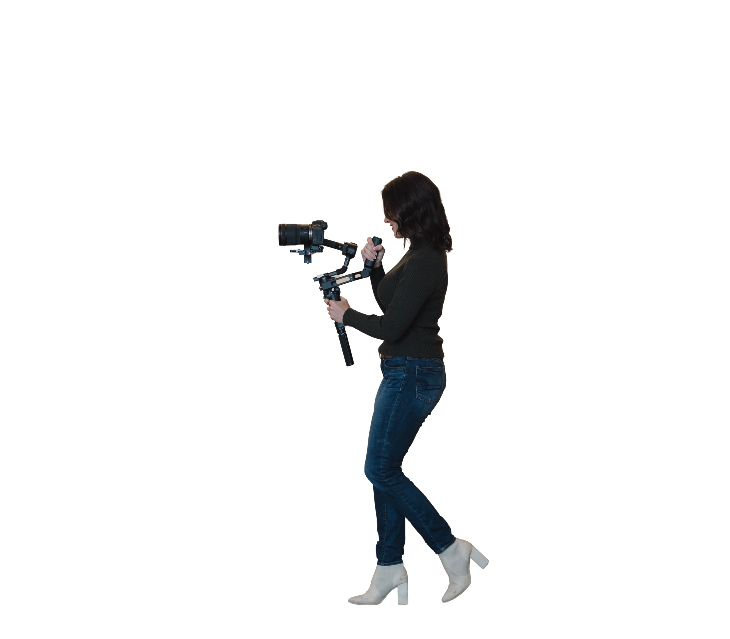 A person with dark hair, wearing a dark sweater, jeans, and white boots, walks while holding a camera mounted on a handheld stabilizer against a white background—capturing dynamic moments that could be used in a professional headshot or during an on-location photo shoot.