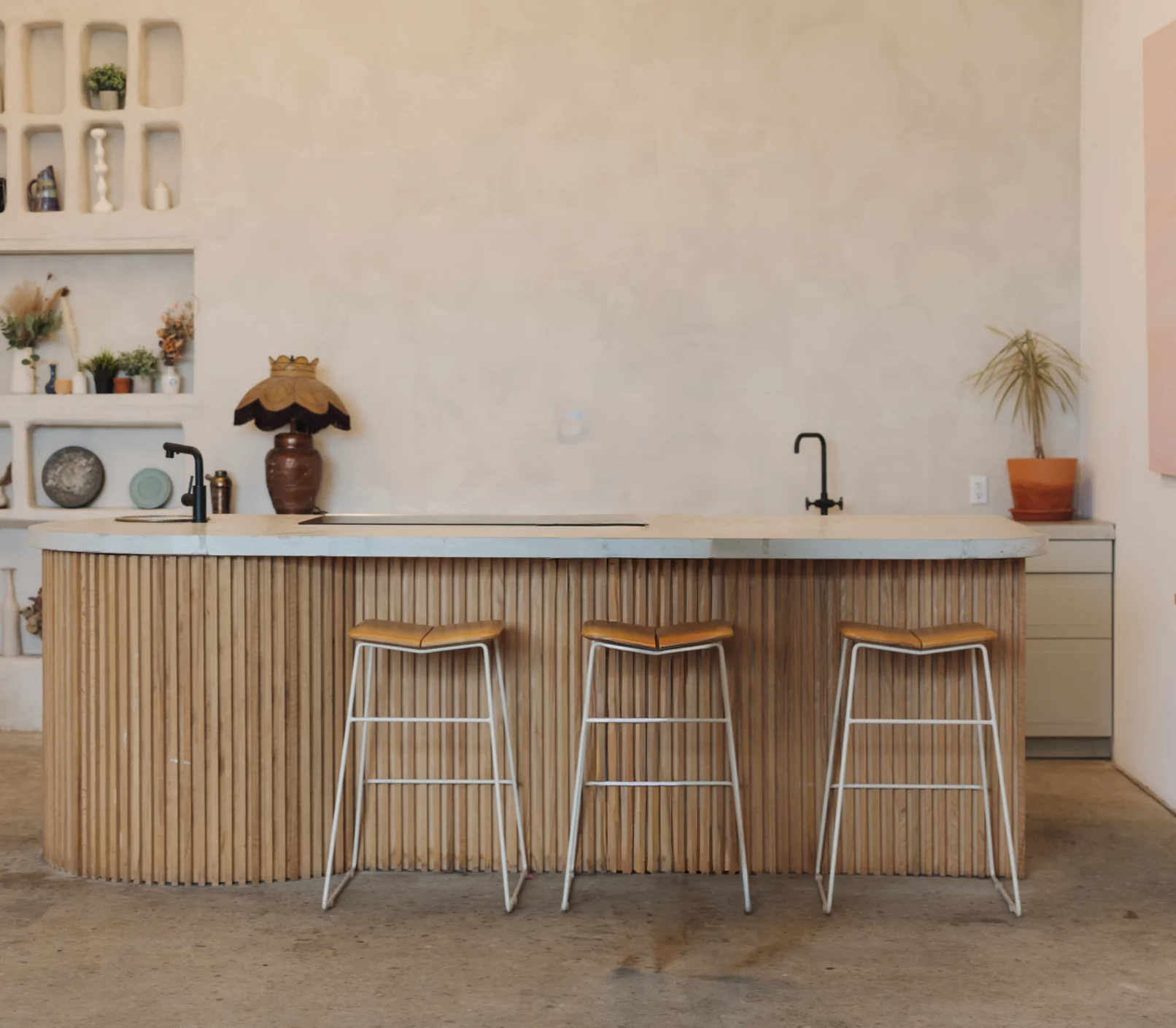 Modern kitchen with a curved wooden island, three bar stools, two black faucets, open shelves displaying decor, and neutral tones throughout. Plants and pottery add a touch of style to the minimalist space—perfectly highlighted through real estate photography or enhanced by virtual staging for an elevated presentation.