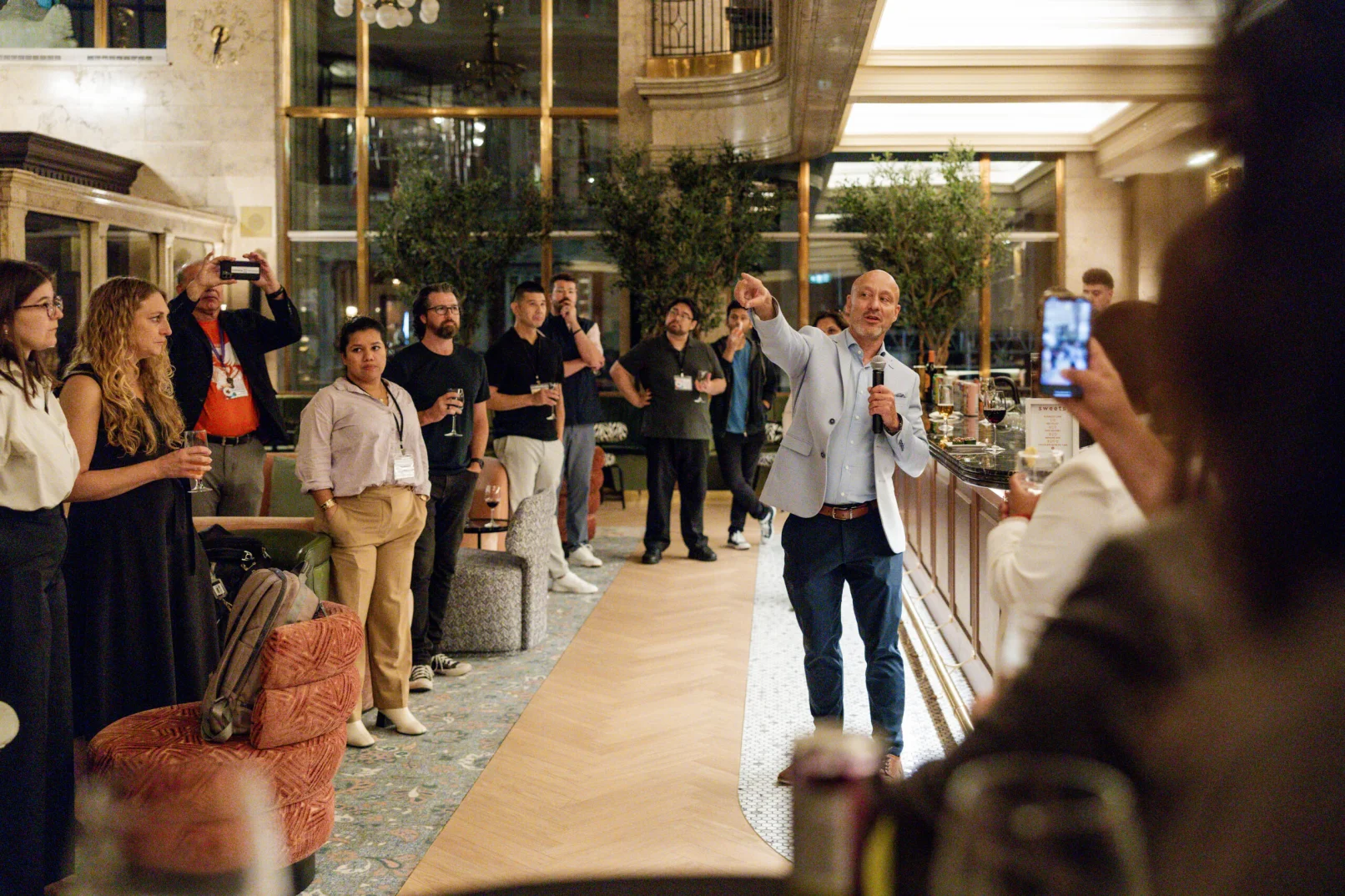 A man speaks to a group of people in a stylish indoor venue, captured through expert event photography. Some attendees listen while others take photos or hold drinks. The setting features elegant decor and soft lighting, creating an inviting atmosphere for memorable moments.