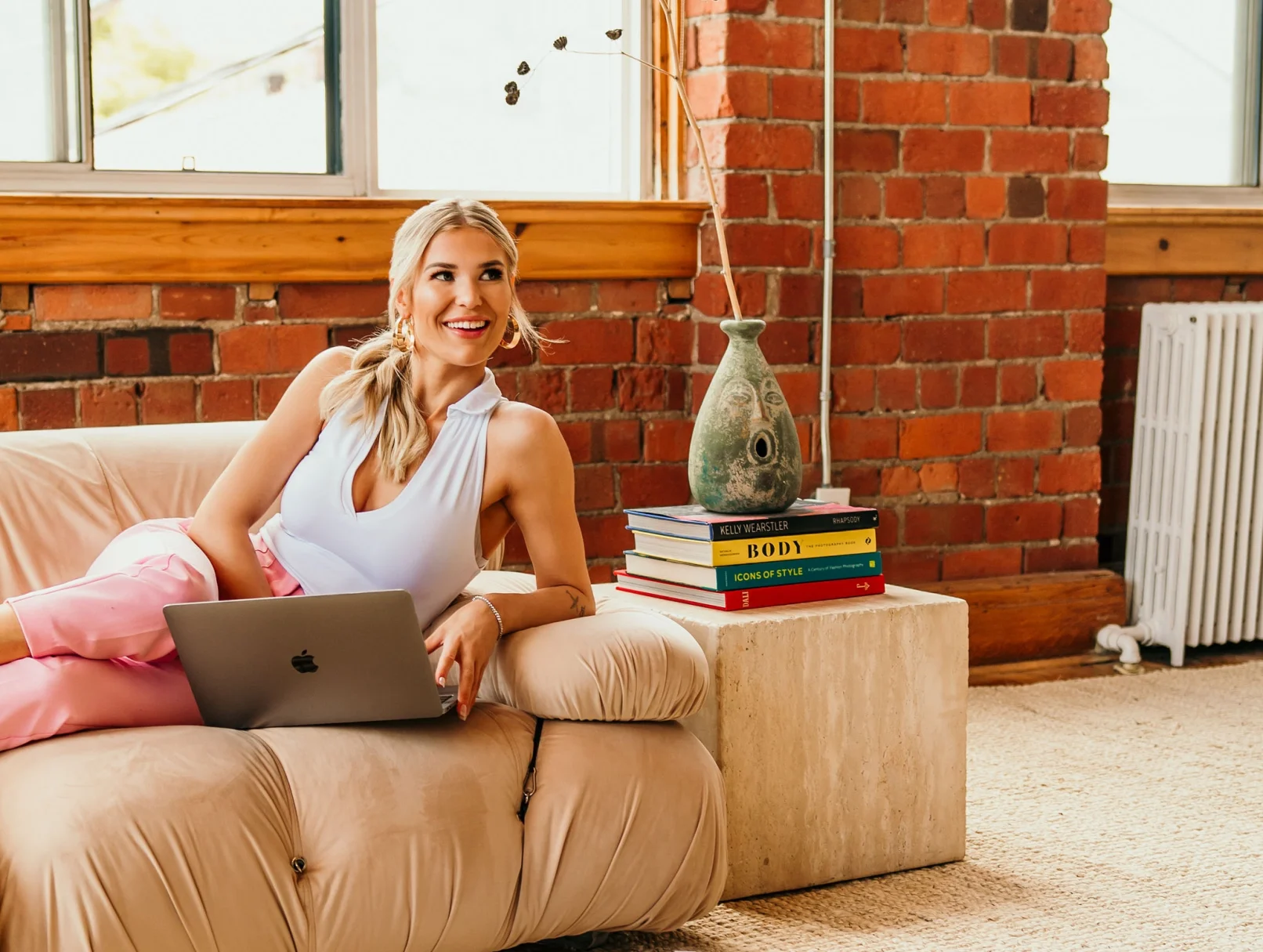 A woman in pink pants and a white top lounges on a beige sofa with a laptop, her smile radiating confidence—perfect for professional headshots. Next to her is a stack of books and a decorative vase on a stone table, set against a brick wall and large window, creating an inviting atmosphere ideal for virtual staging.